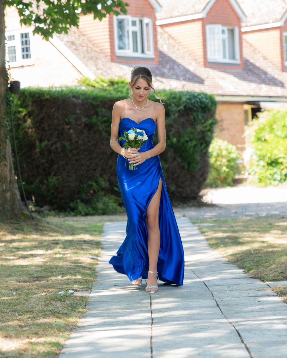 Bridesmaid in royal blue satin strapless gown holding white bouquet walking down pathway at outdoor wedding.