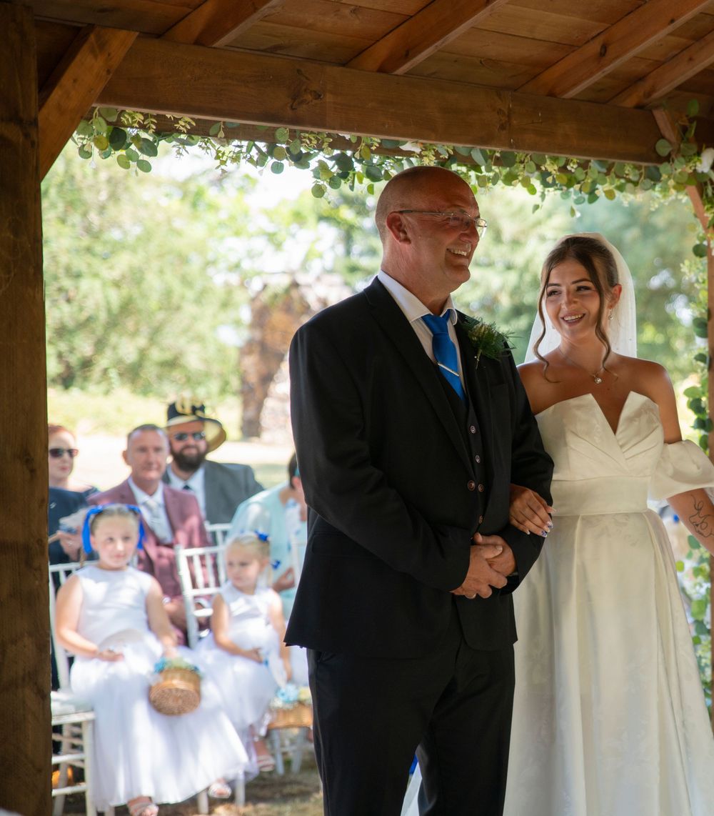 Smiling bride and groom holding hands during outdoor wedding ceremony under wooden pergola with greenery.