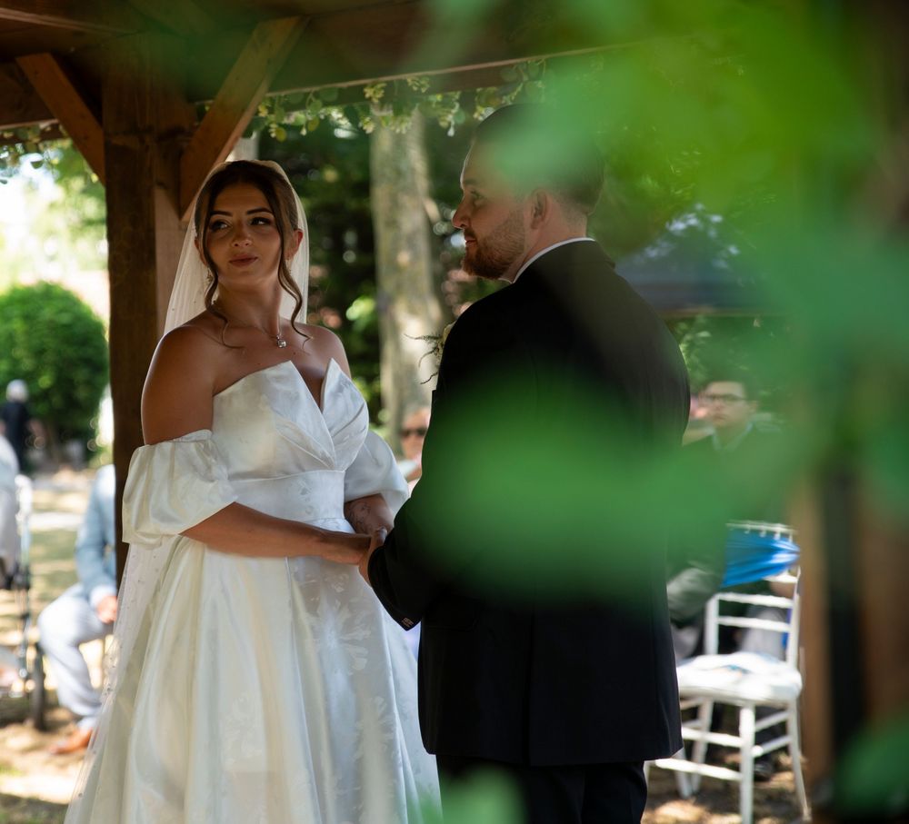 Bride in off-shoulder white gown exchanges vows with groom at outdoor wedding ceremony under a wooden pergola.