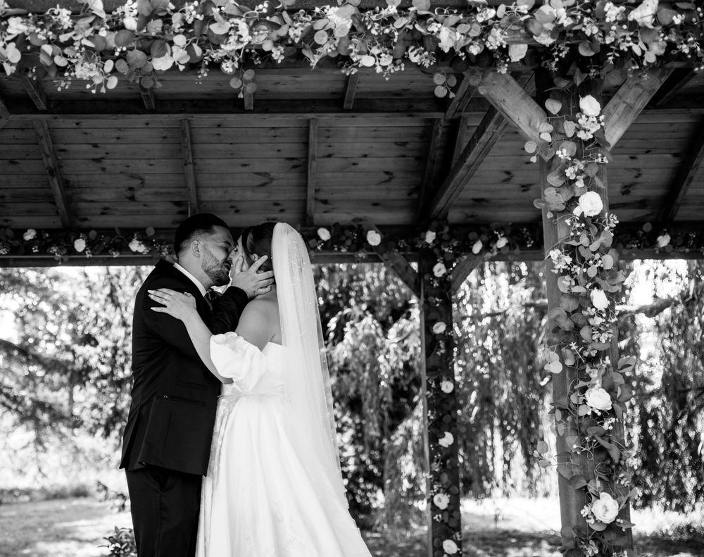 Bride and groom share first kiss under floral arch at outdoor wedding ceremony in black and white.