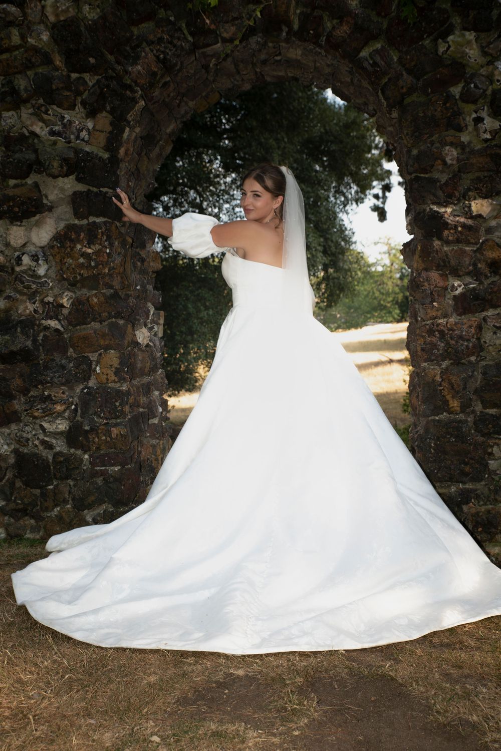 Bride in white ball gown with veil poses gracefully under a rustic stone arch in an outdoor garden setting.