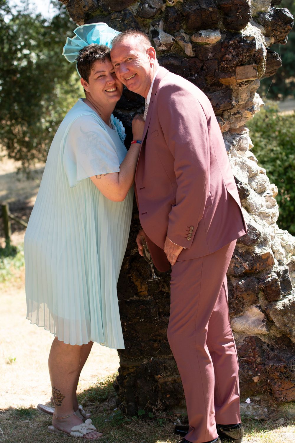 Couple posing by stone pillar outdoors, woman in light blue dress with bow, man in pink suit, smiling.