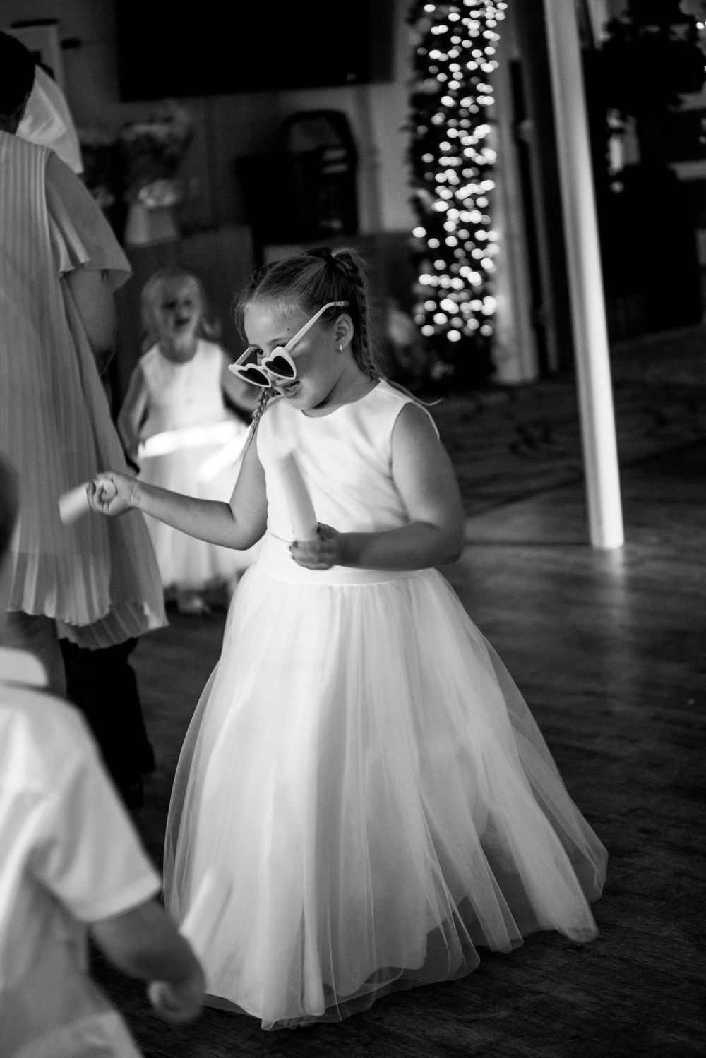 A young flower girl in a white tulle dress and heart-shaped sunglasses dances joyfully at a wedding reception.