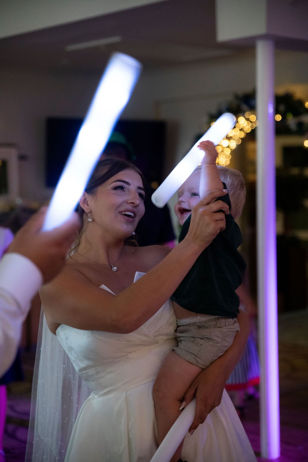 Bride in white gown holding a child with glowing LED foam sticks at a wedding reception dance floor.