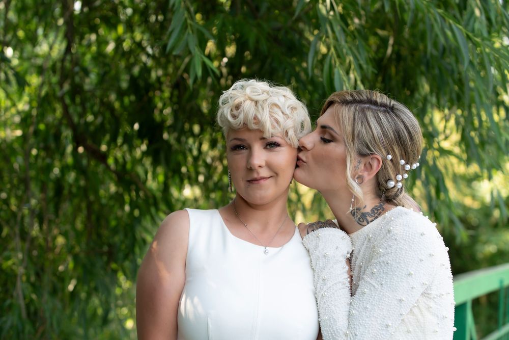 Two brides share a tender kiss at their outdoor wedding ceremony surrounded by lush green trees.