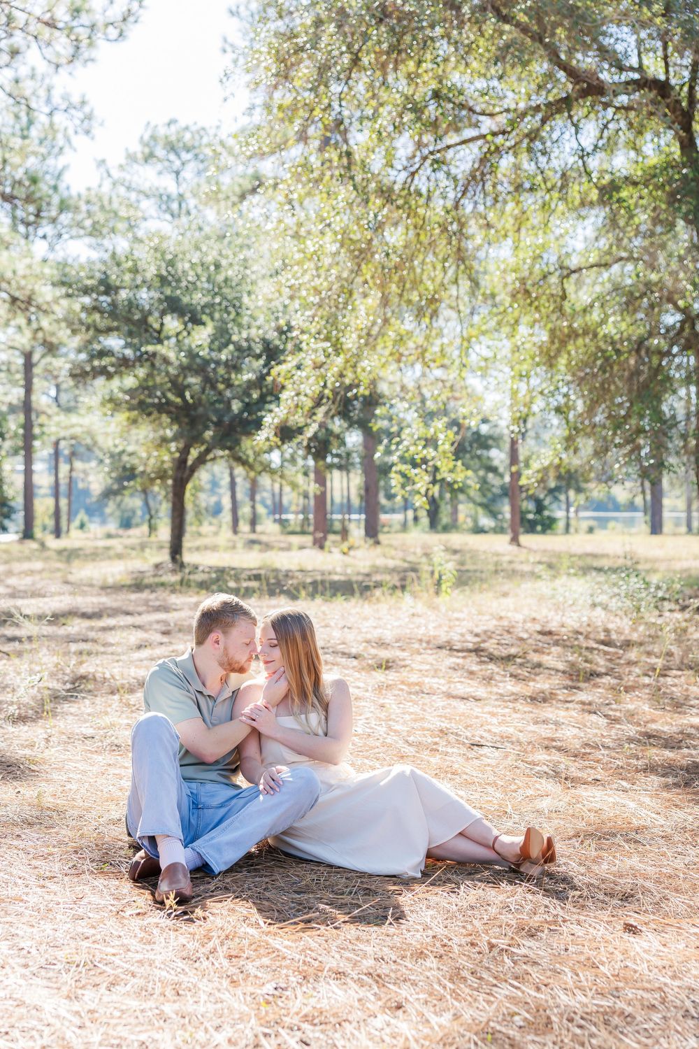 A couple relaxes together on a pine needle covered ground beneath tall trees in a romantic outdoor setting.