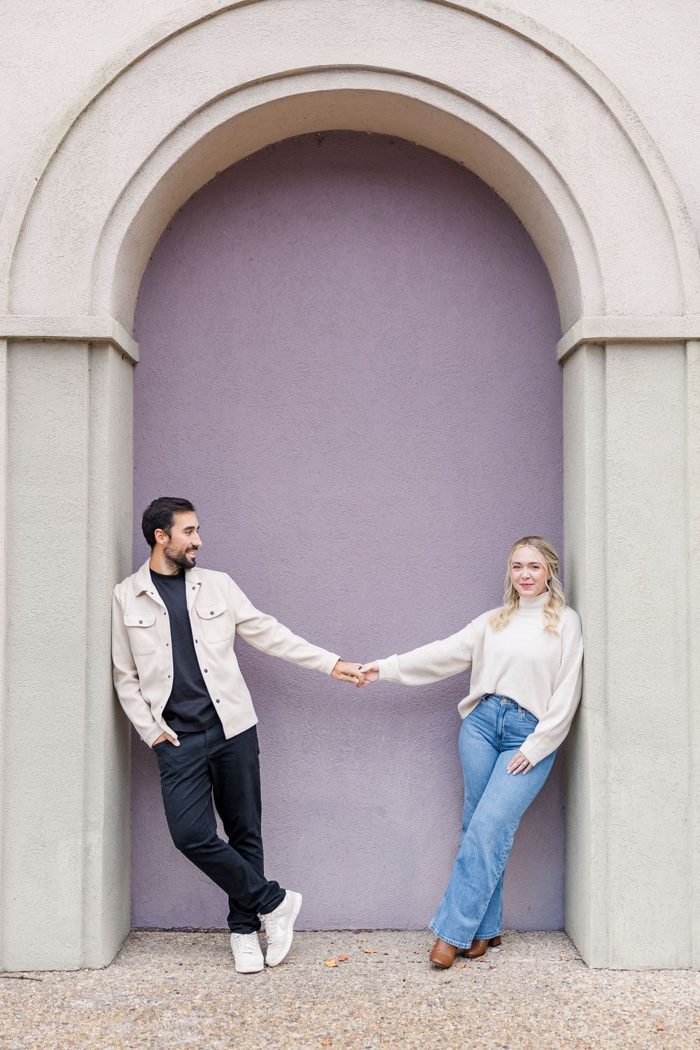 A couple poses playfully in an arched doorway against a purple wall, holding hands while leaning in opposite directions.