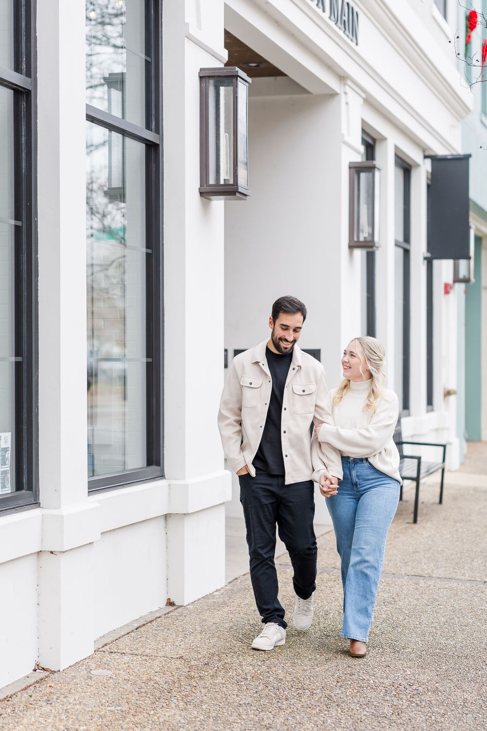 A couple walks and laughs together along a white building exterior with large windows and textured ground.