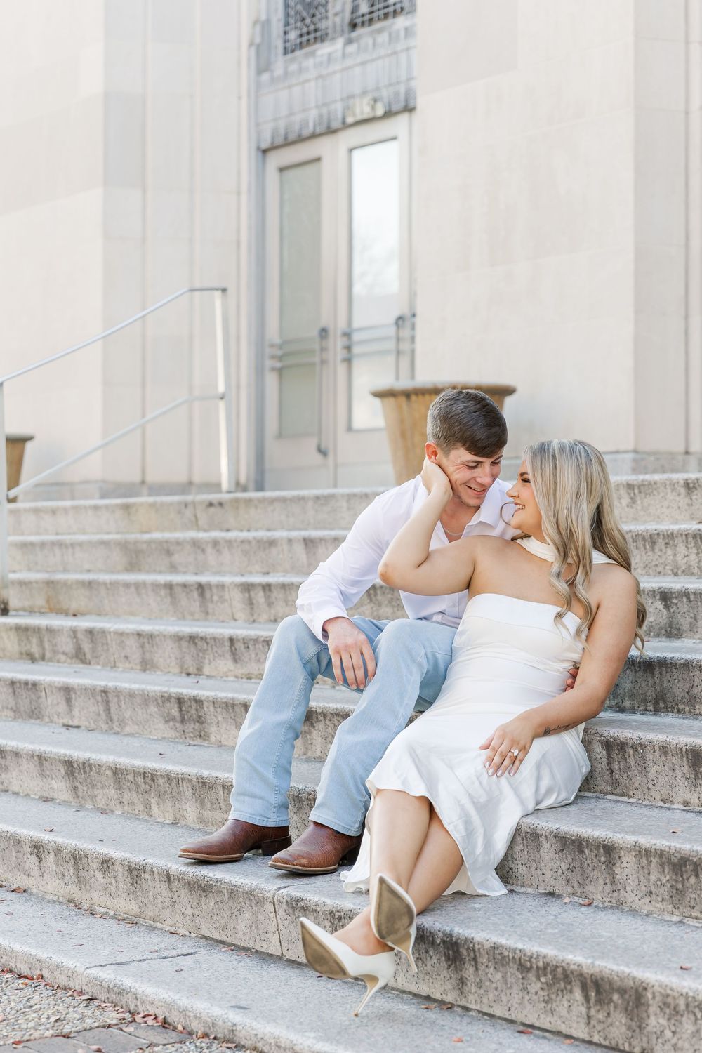 A couple in white and light blue outfits sits intimately on concrete steps during an outdoor photo session.
