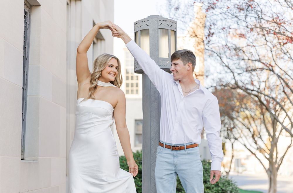 Couple in white outfits poses together during outdoor engagement photo session near white building.