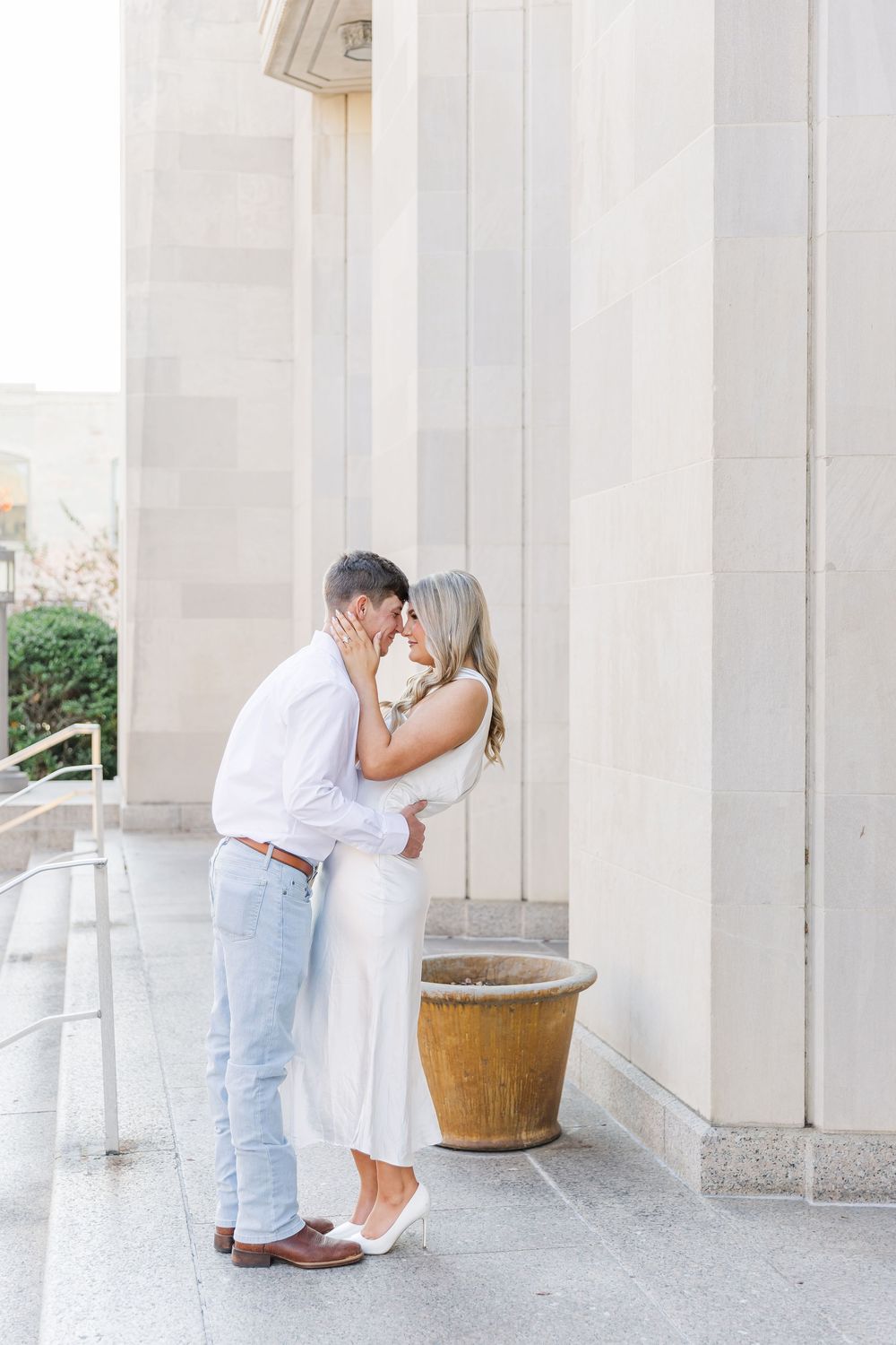 A romantic couple sharing an intimate moment between white stone columns on a bright sunny day.