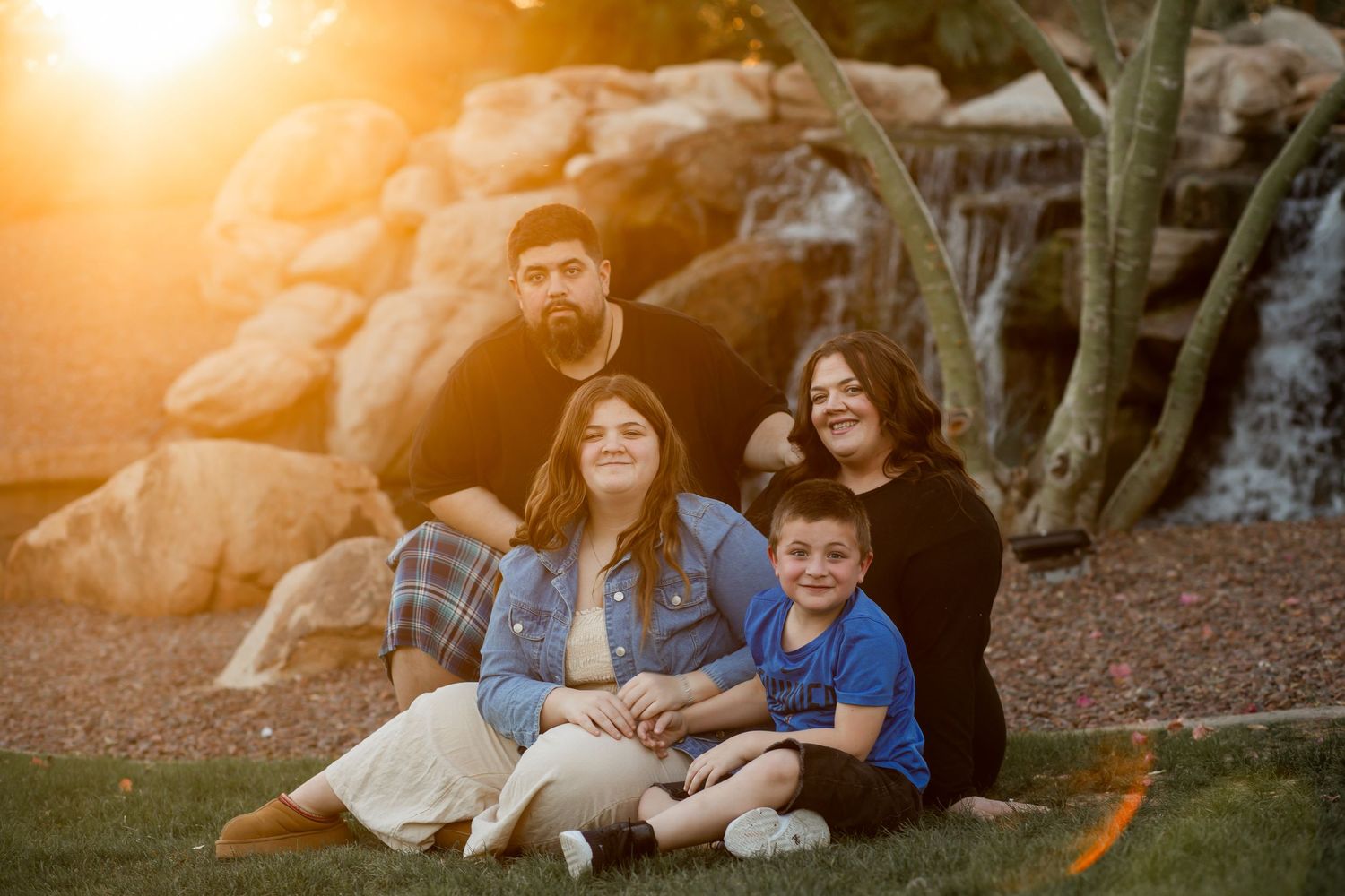 Family of four smiles together on grass at sunset near rocky waterfall backdrop.