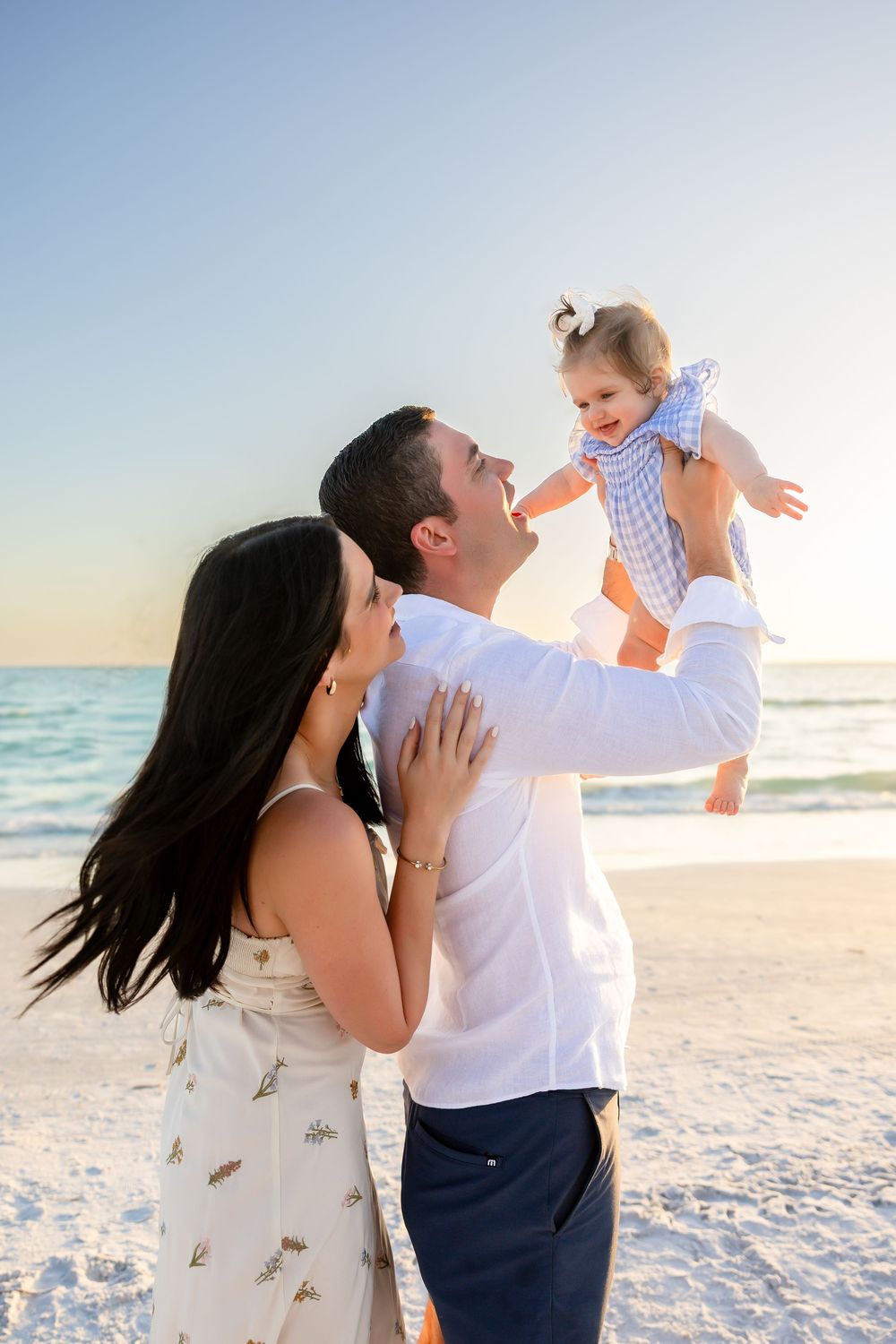Happy couple lifts laughing baby girl on sunny beach at golden hour.