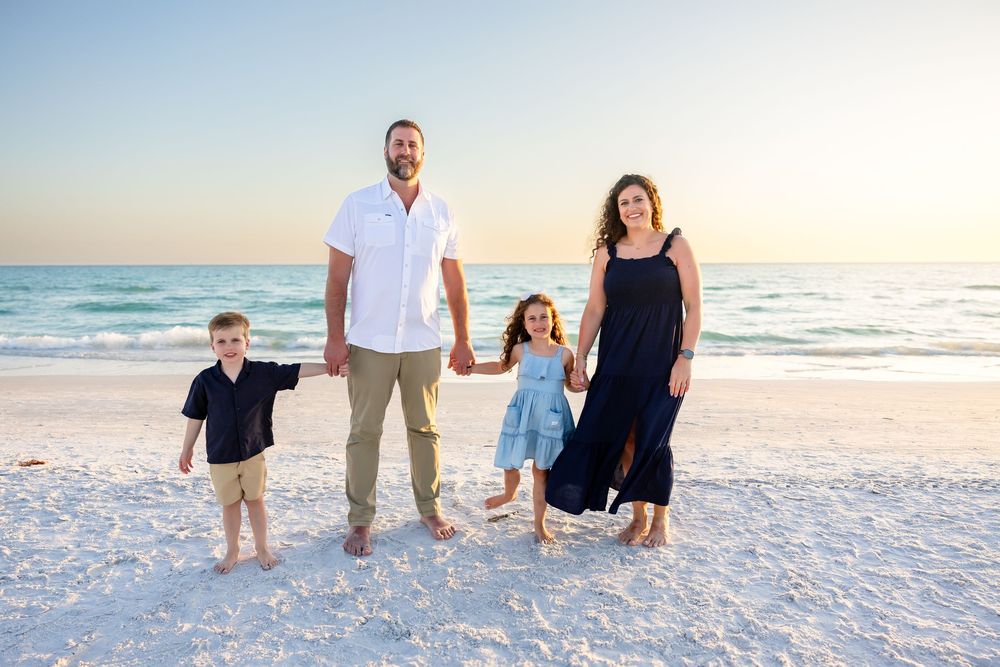 Family of four holds hands smiling on white sand beach at sunset.