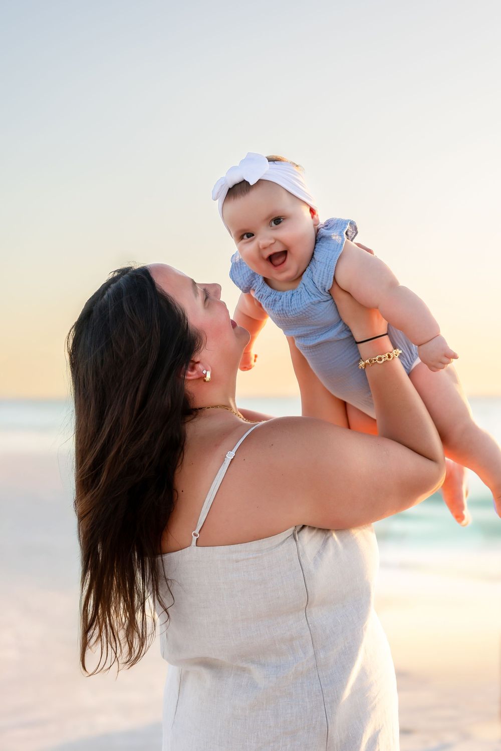 Mother lifts smiling baby girl with white bow on sunny beach.