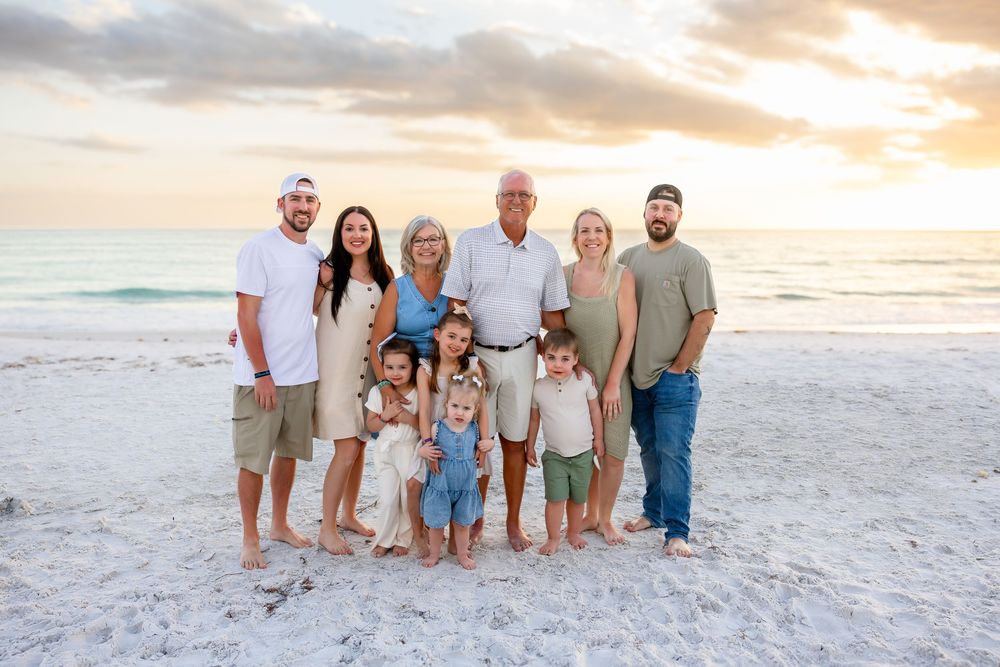 Multi-generational family of ten posing together on a white sand beach during a colorful sunset.