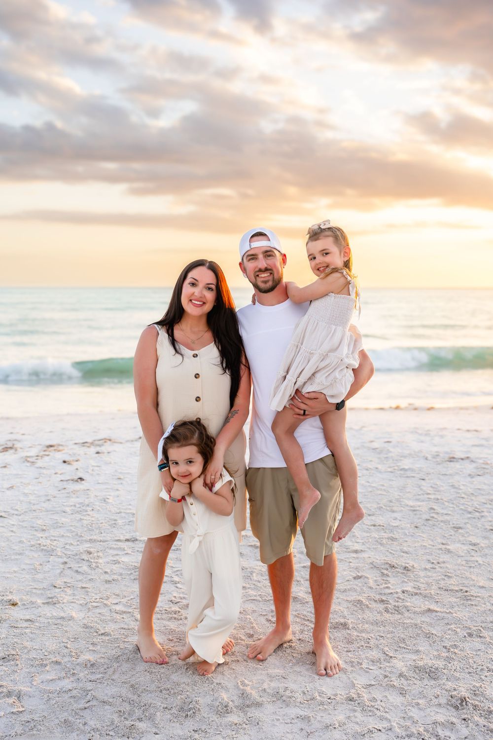 Family of four posing on a sandy beach at sunset, father holding daughter while toddler stands in front.
