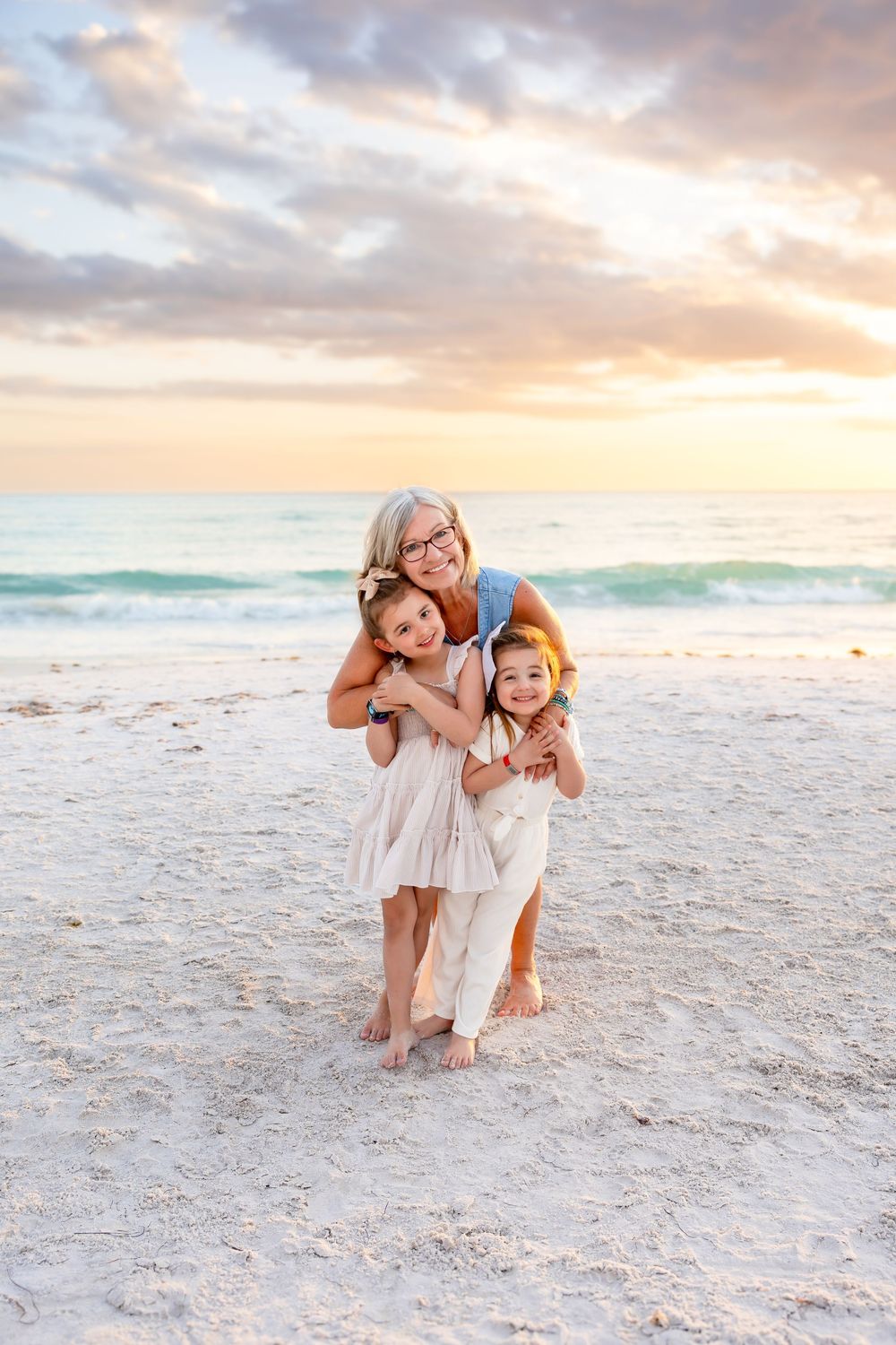 Grandmother hugging two young grandchildren on a white sand beach during a pastel sunset.