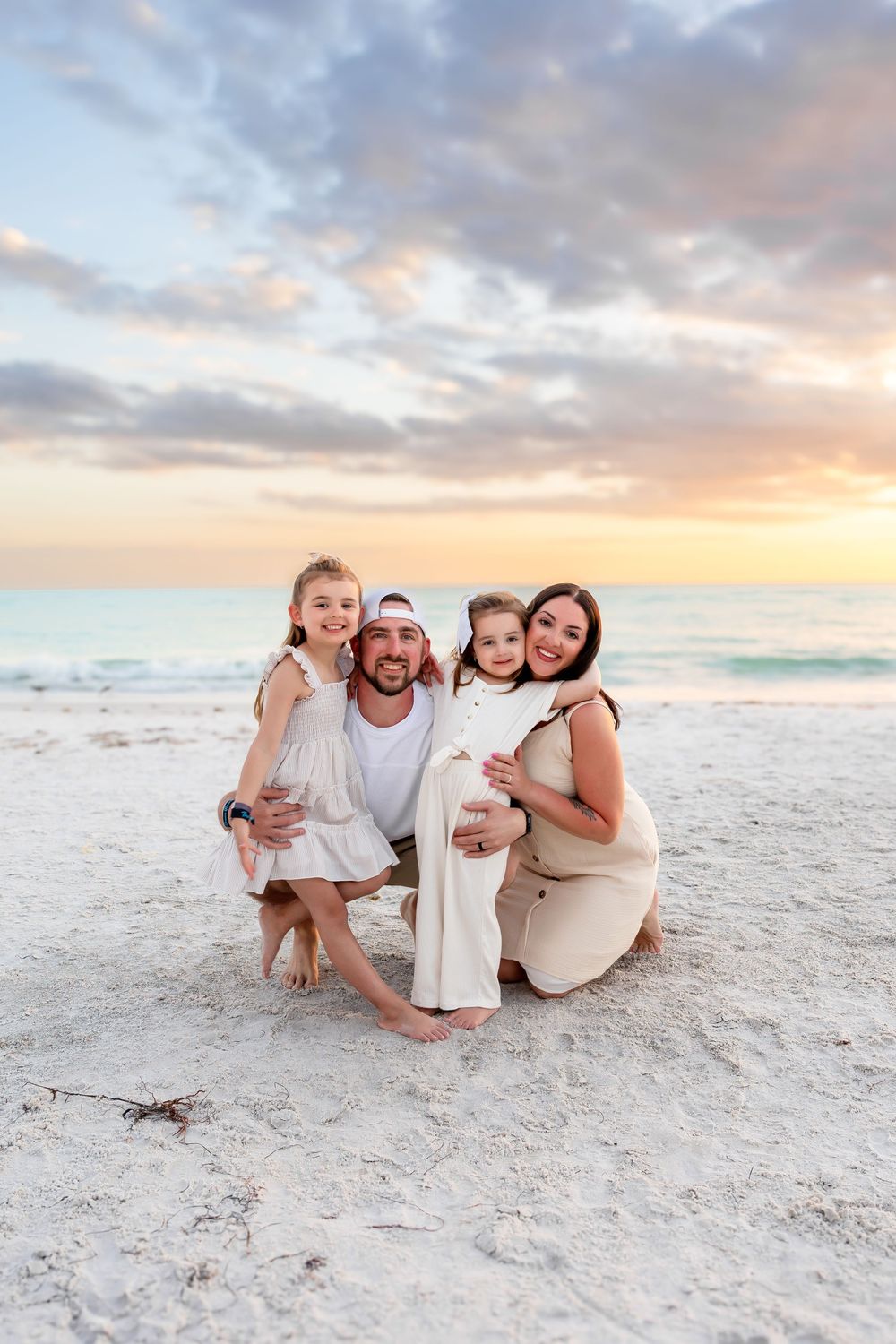 Young family of four crouching together on a sandy beach smiling during a vibrant sunset.