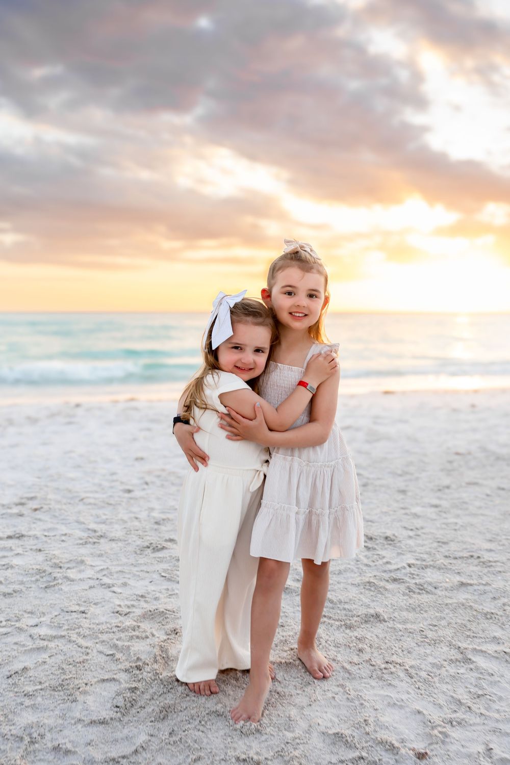 Two young sisters hugging on a white sand beach at sunset, wearing matching cream dresses.
