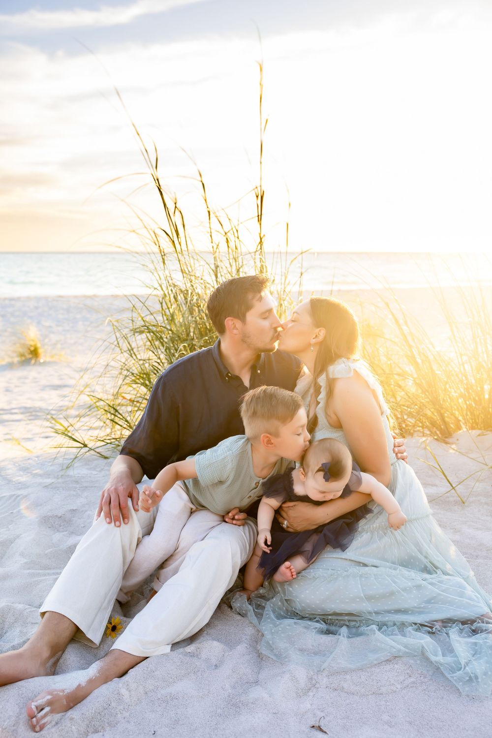 Family sitting on beach sand at sunset, parents kissing while two young children sit between them near sea grass.