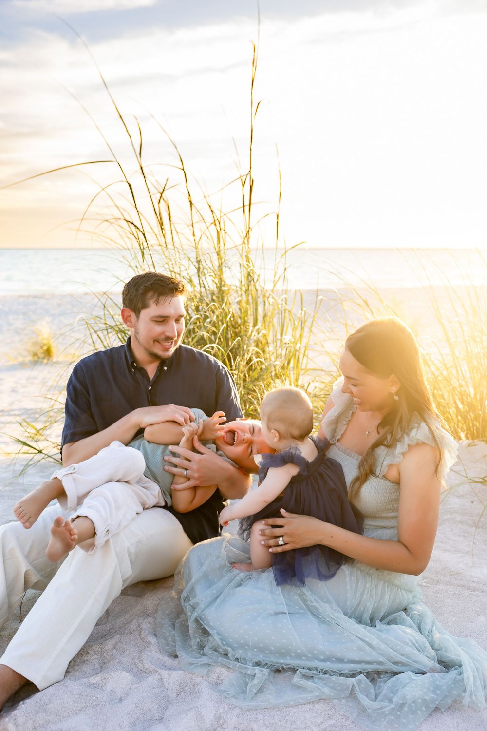 Family sitting on sandy beach at sunset, parents laughing with baby, sea oats and ocean in background.