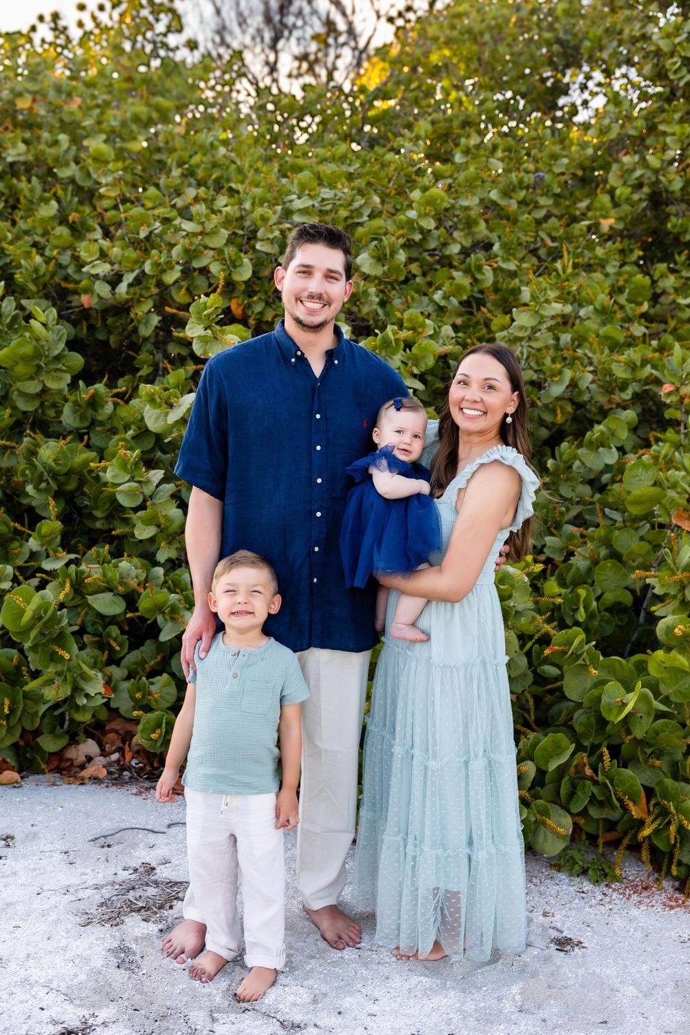 A happy family of four poses outdoors near lush green shrubs on a sandy beach, dressed in coordinating blue and green outfits.