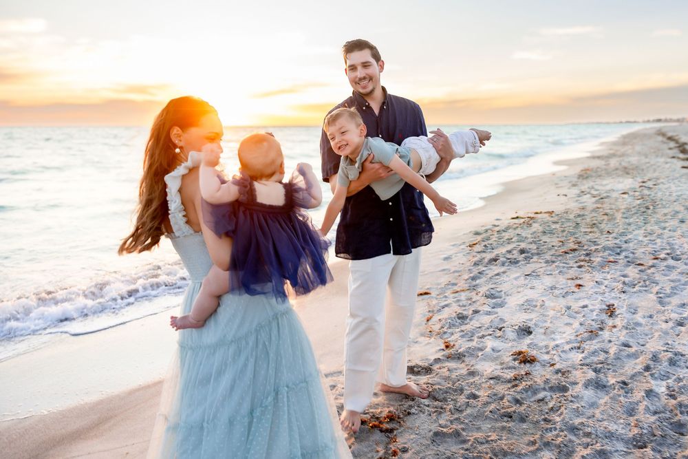 Family of four enjoying a golden sunset beach walk, parents carrying toddlers in blue and white outfits on sandy shore.