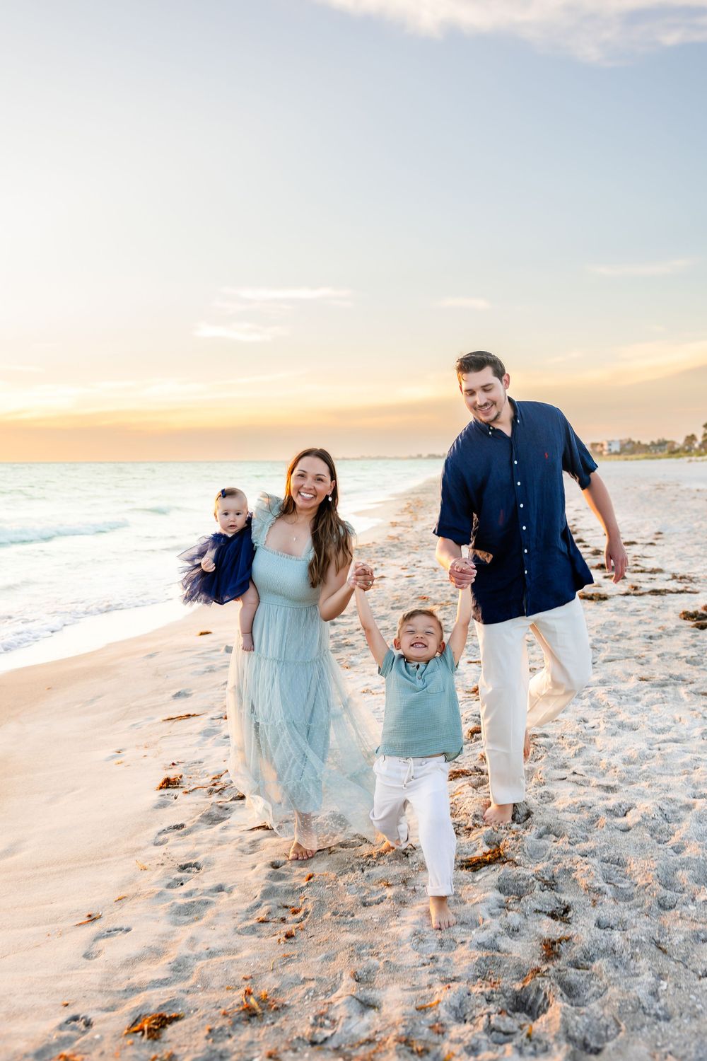 Happy family of four enjoying a sunset beach walk, parents swinging toddler son while mom holds baby girl.