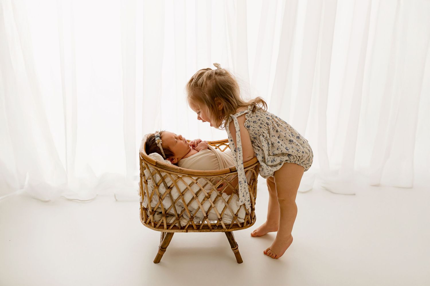 Toddler sister peeking into rattan bassinet at newborn baby during sibling portrait session at Flowertown Photography studio in Summerville