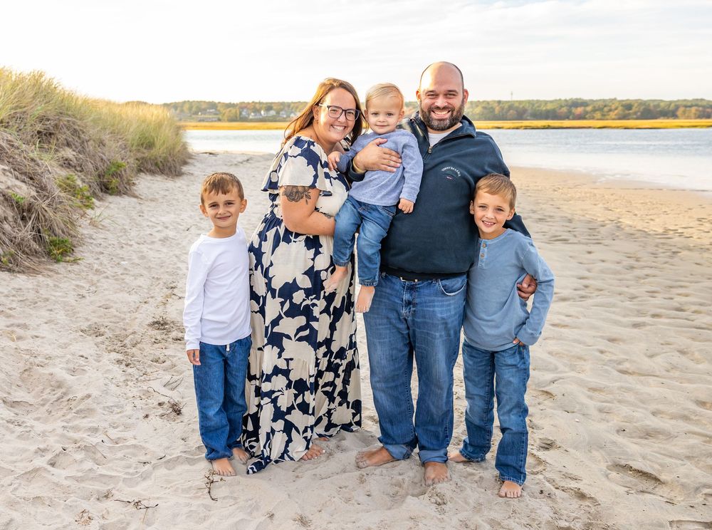 A group stands together smiling on a sandy beach with water and autumn foliage in the background.