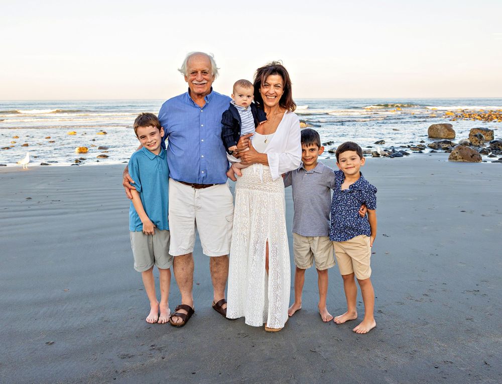 Family in coordinated blue and white outfits poses together on a beach at sunset with ocean waves in background.