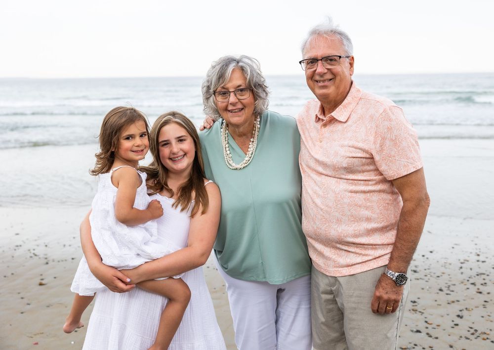 A multi-generational family poses together smiling on a sunny beach with ocean waves in the background.