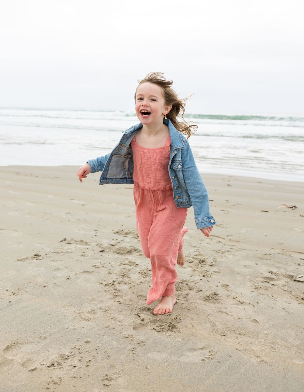 Young child in a coral dress and denim jacket runs joyfully along a misty beach shoreline.