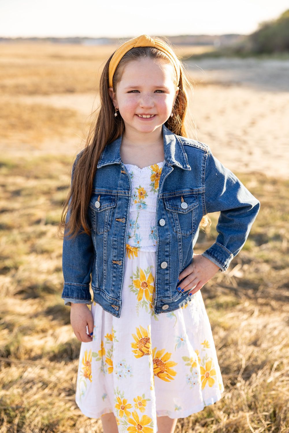 A young child in a denim jacket and floral dress poses in golden sunlight during sunset.
