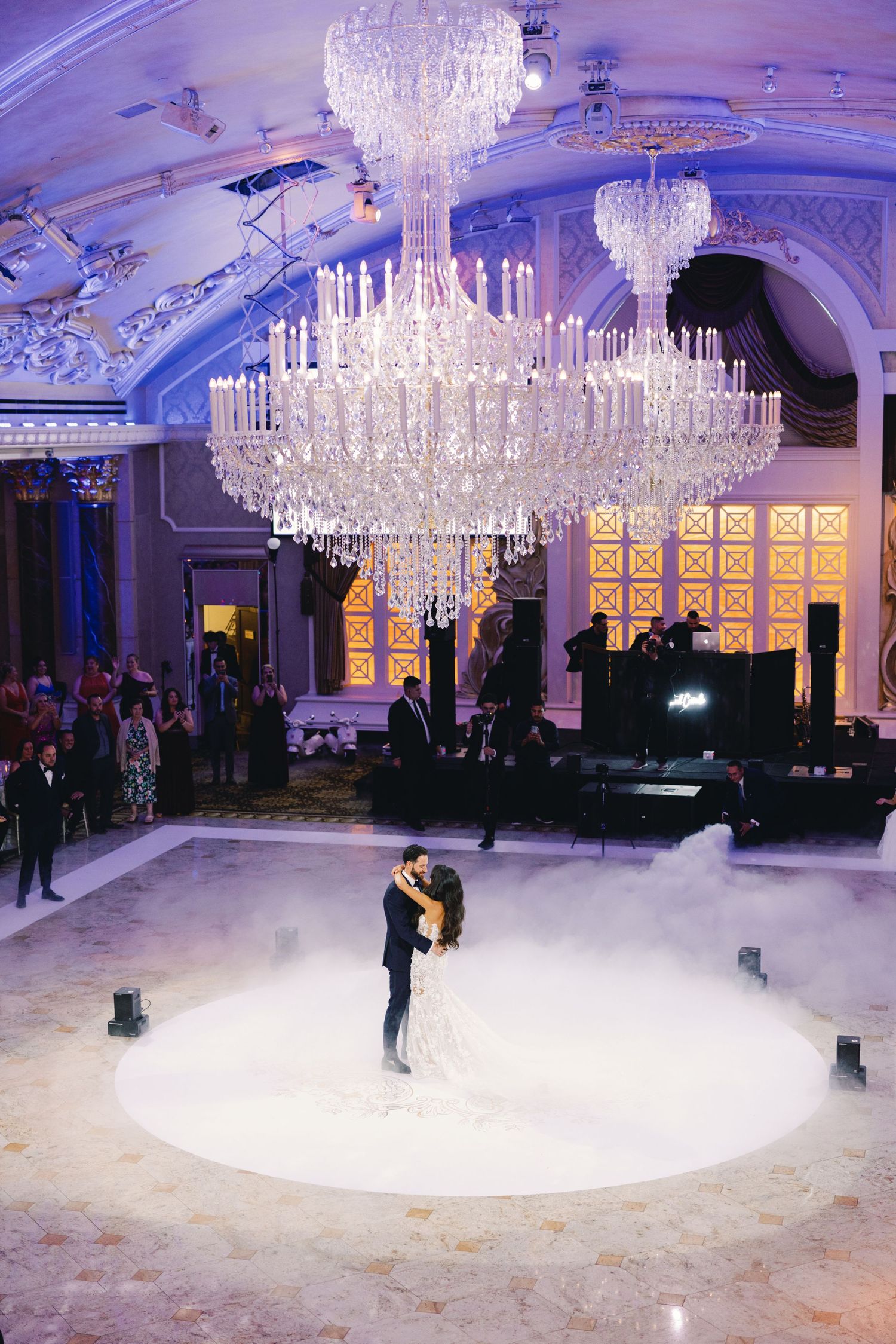Bride and groom share first dance under grand crystal chandeliers with dramatic fog on dance floor.