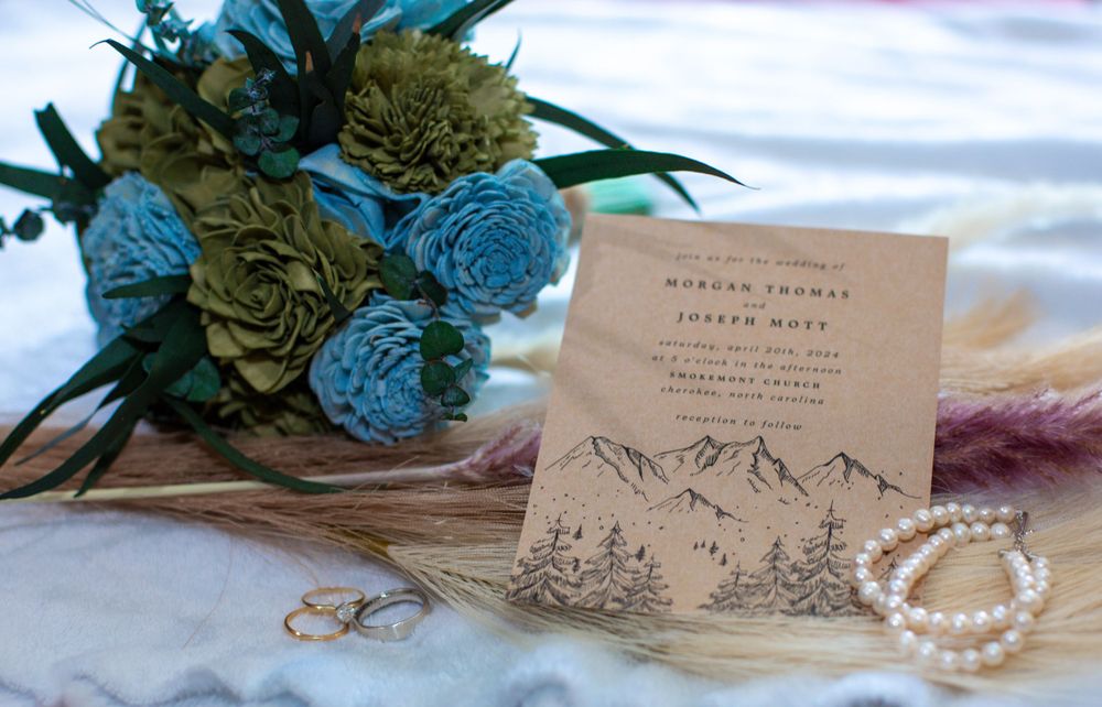 Wedding details featuring blue flowers, mountain-themed invitation, rings and pearl bracelet.