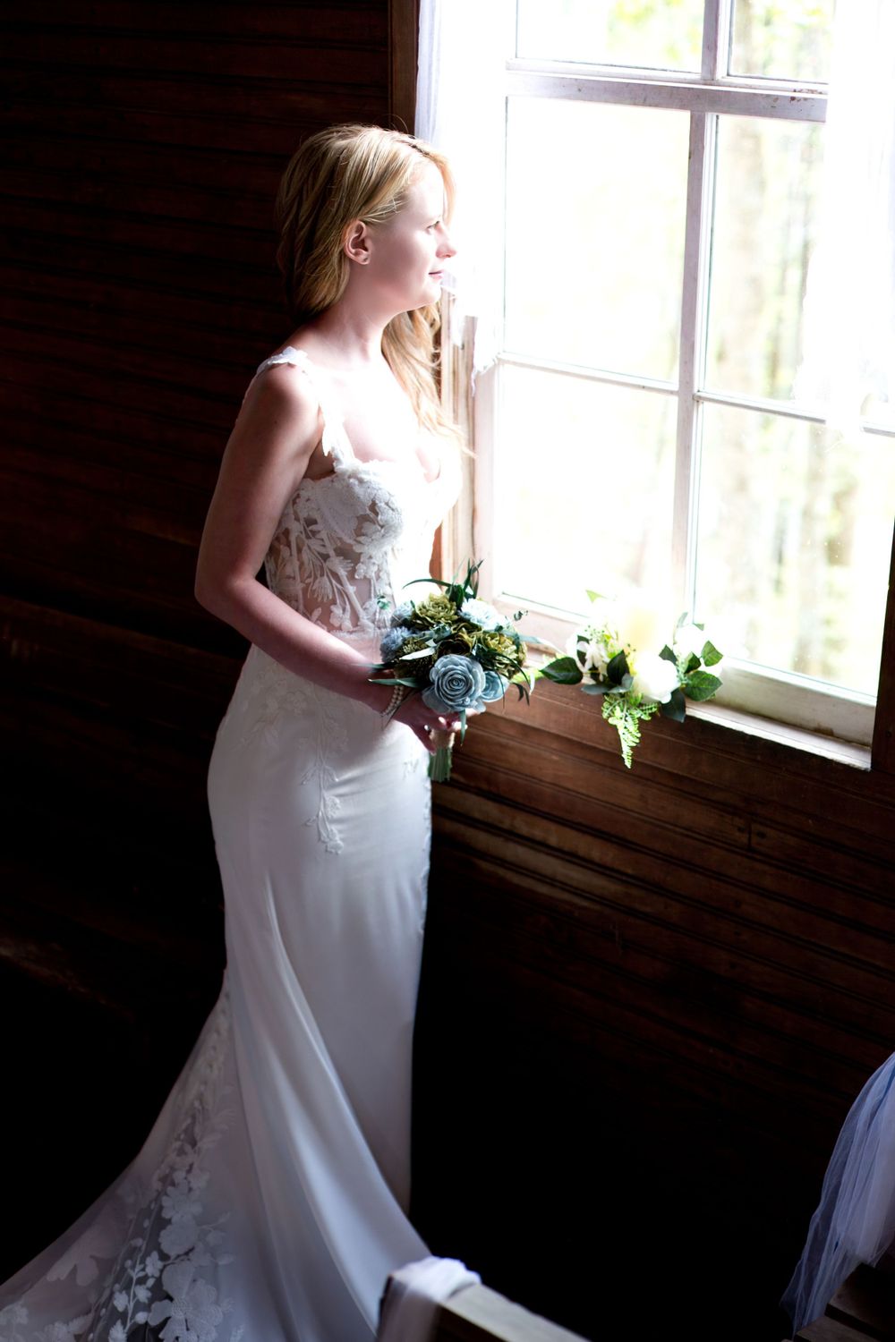 An elegant white wedding gown silhouetted against a bright window with a floral bouquet.