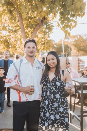 A couple stands together at an outdoor event during golden hour with autumn leaves in the background.