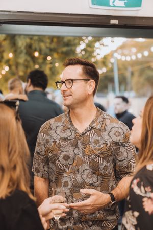 A person wearing glasses and a patterned shirt mingles at an indoor social gathering with string lights in the background.