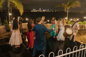 Guests dancing outdoors at a nighttime waterfront celebration with palm trees and city lights in the background.