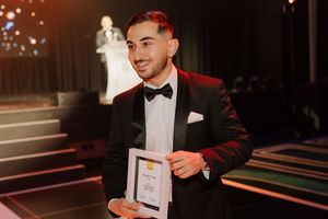 Young man in tuxedo holding award certificate at formal black tie gala event.