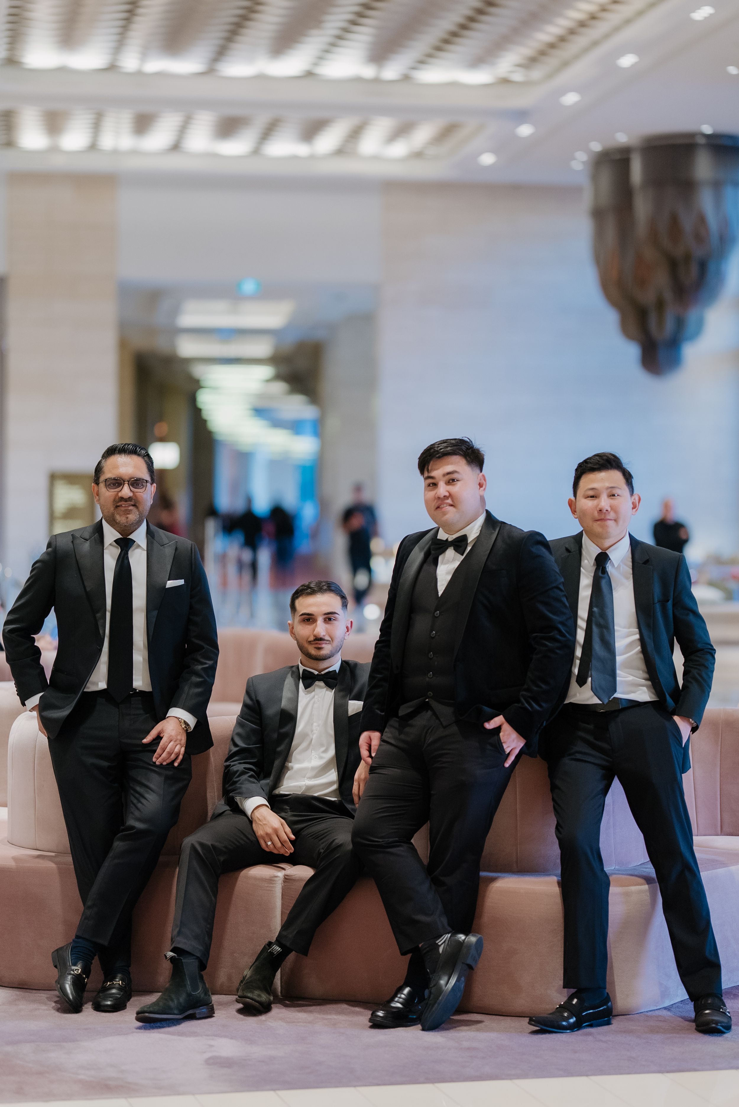 Four men in formal black suits pose elegantly in a modern venue lobby for a wedding groomsmen photo.