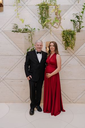 Elderly man in tuxedo and pregnant woman in red gown pose together at formal event with greenery backdrop.