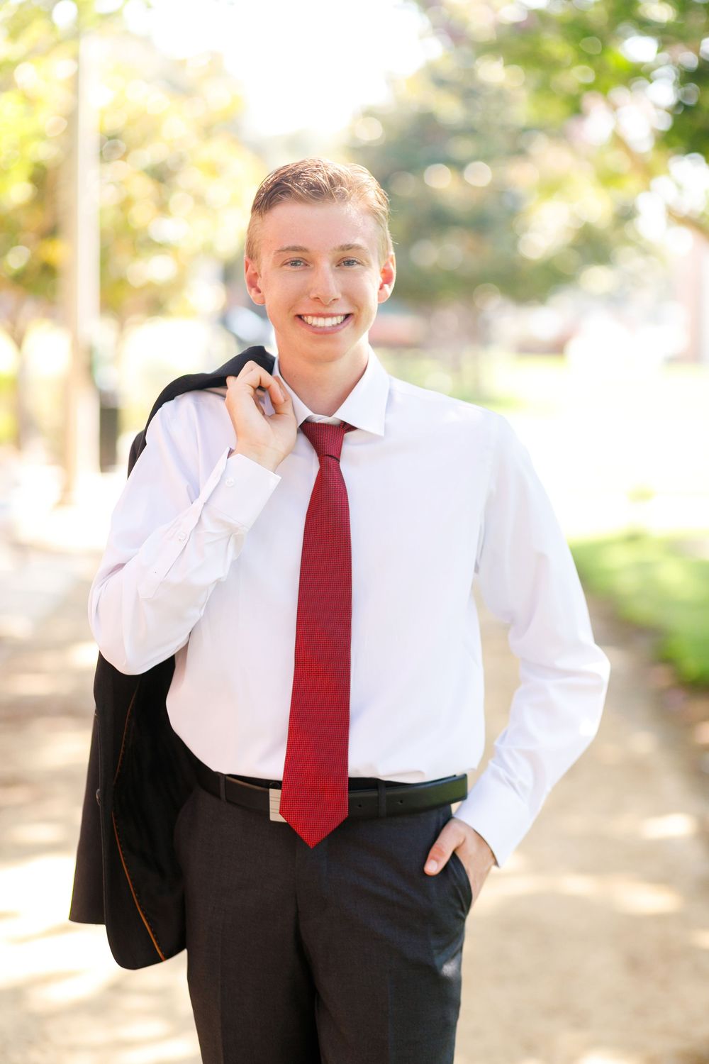 Young professional man in white shirt and red tie smiling outdoors, holding jacket over shoulder.