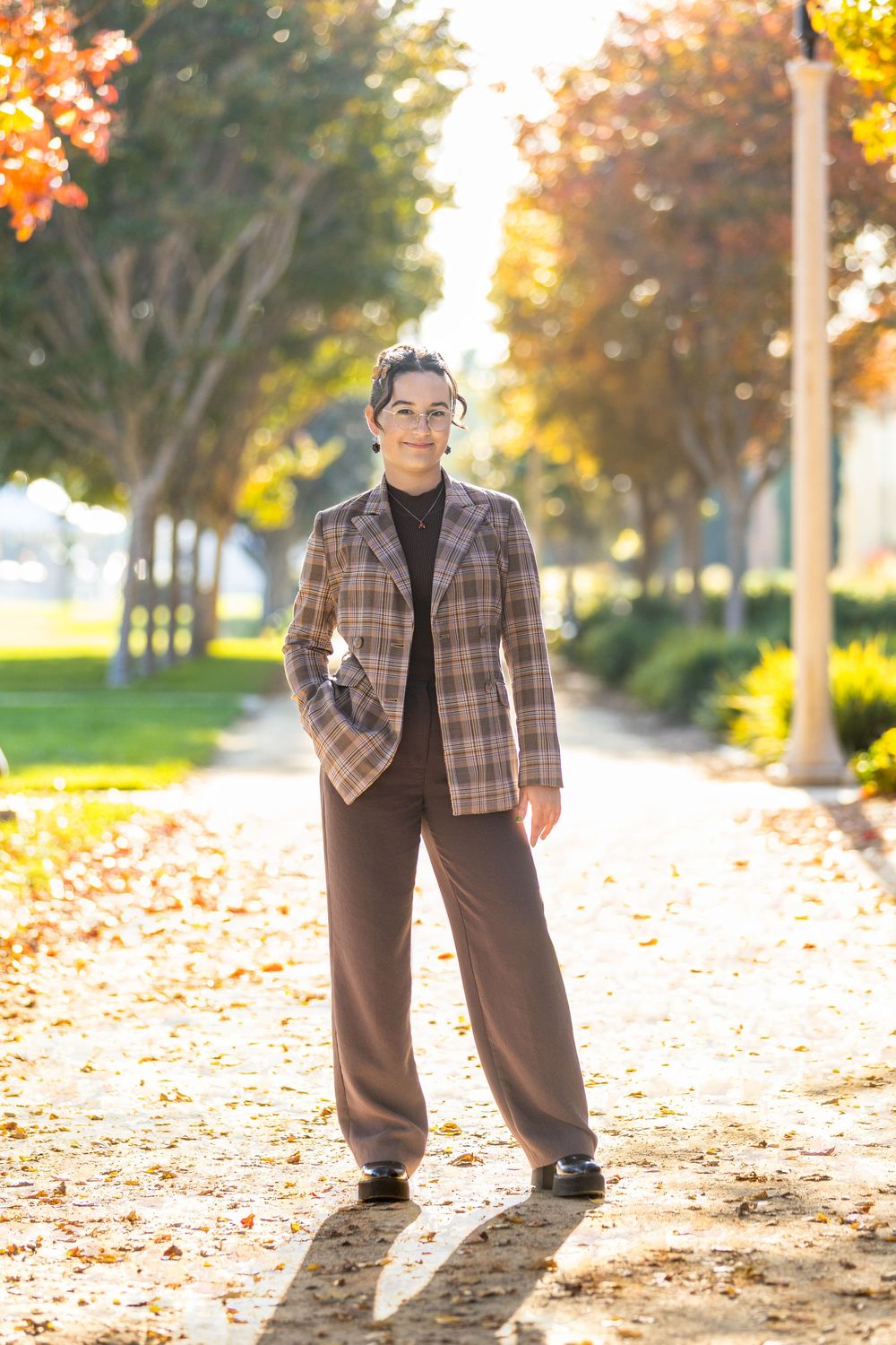 Woman in plaid blazer and brown trousers standing on a sunlit autumn park path with colorful foliage.