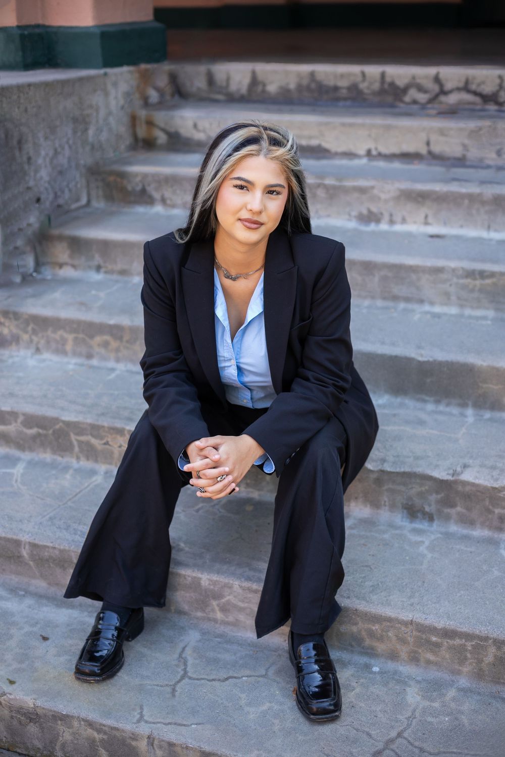 Professional in black suit and blue shirt sitting on concrete steps in an outdoor urban setting.