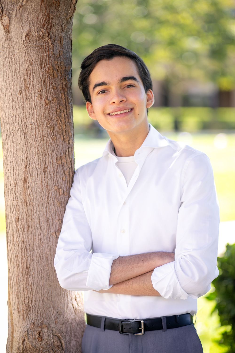 Smiling young man in white shirt leaning against tree with arms crossed outdoors.