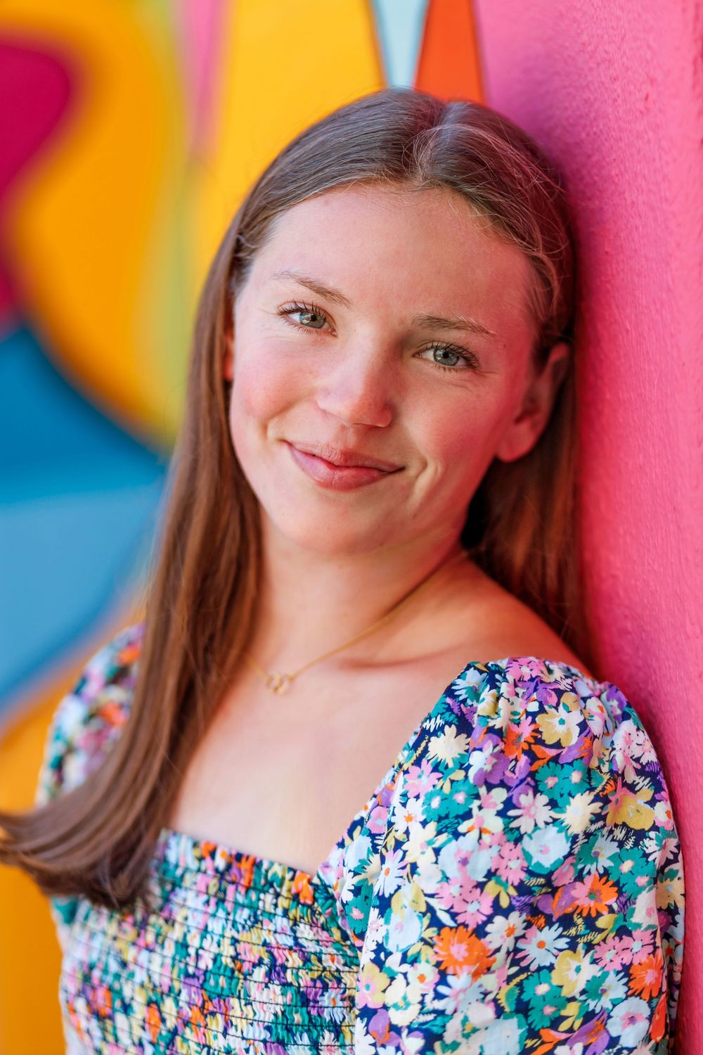 Smiling teenage girl in floral top leaning against colorful painted wall outdoors.