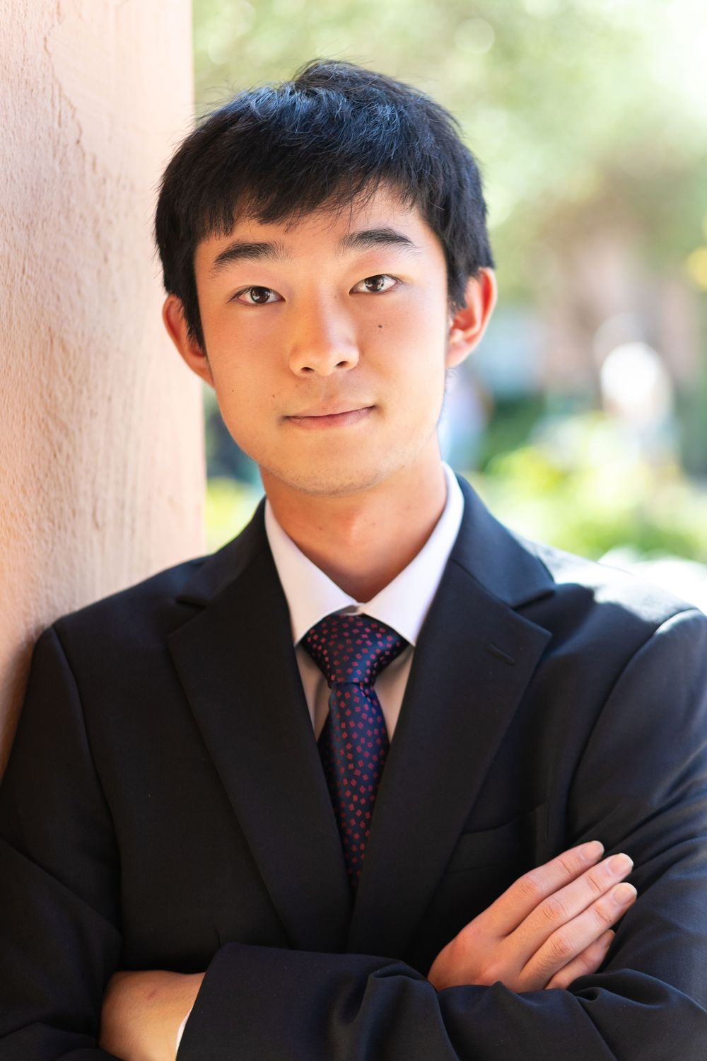 Young Asian man in black suit and purple tie, arms crossed, leaning against a wall outdoors.
